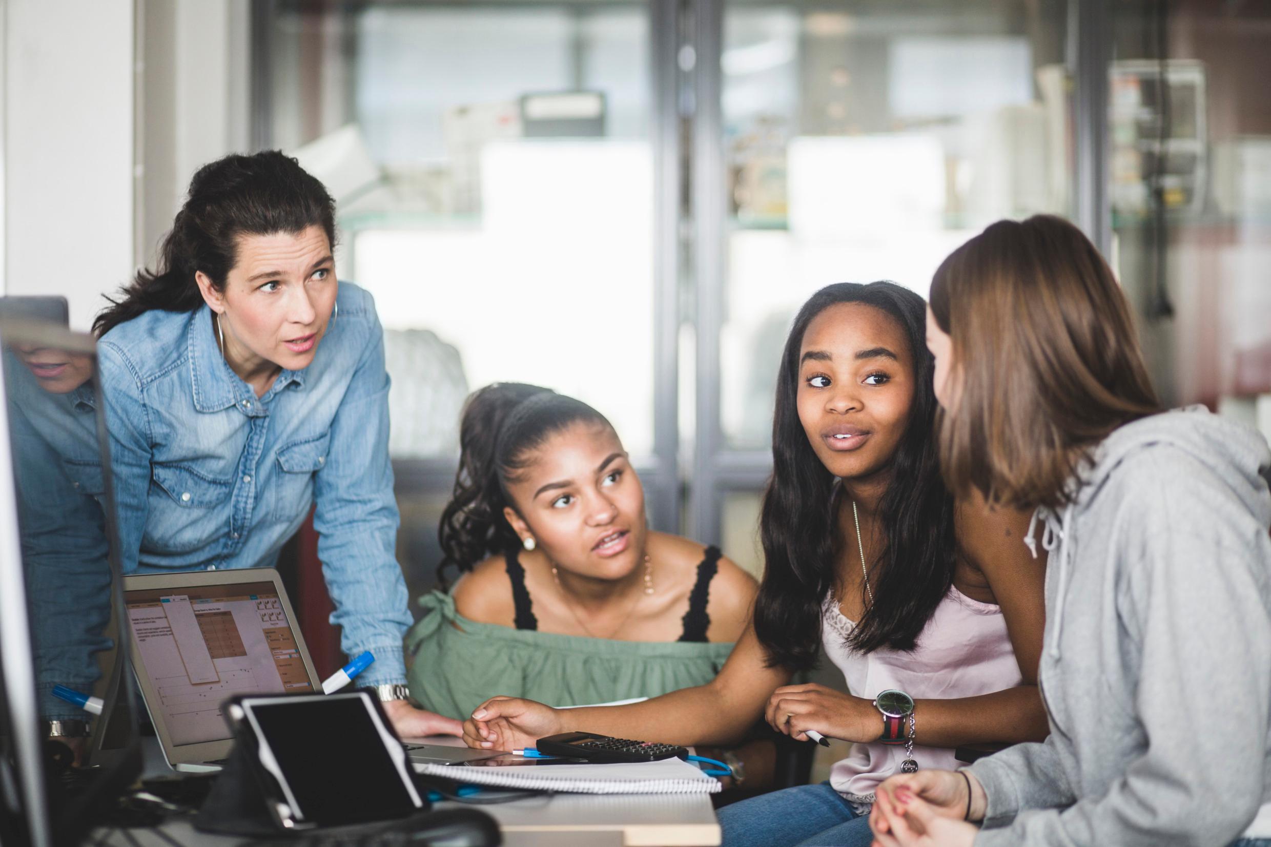 A woman leans against a desk by which three girls are sitting.