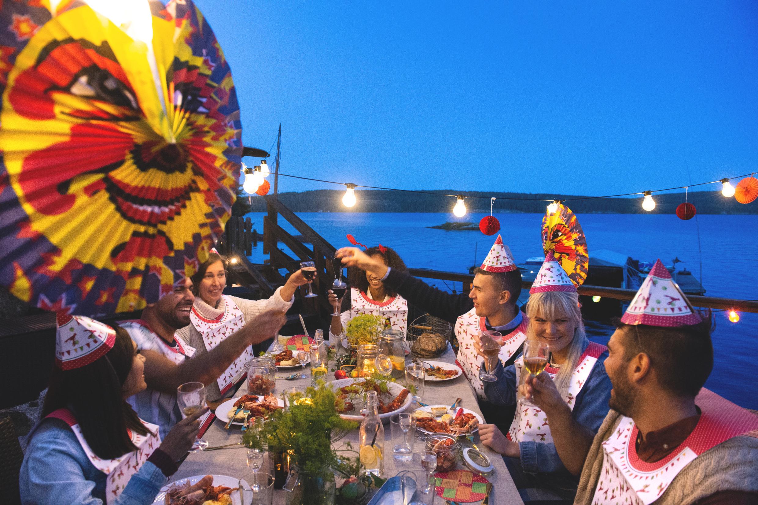 Crayfish party at a jetty by the sea. Seven peoples around a table dressed in paper hats and bibs are eating crayfish and toasting with beer and wine. There are paper lanterns hanging above the table.