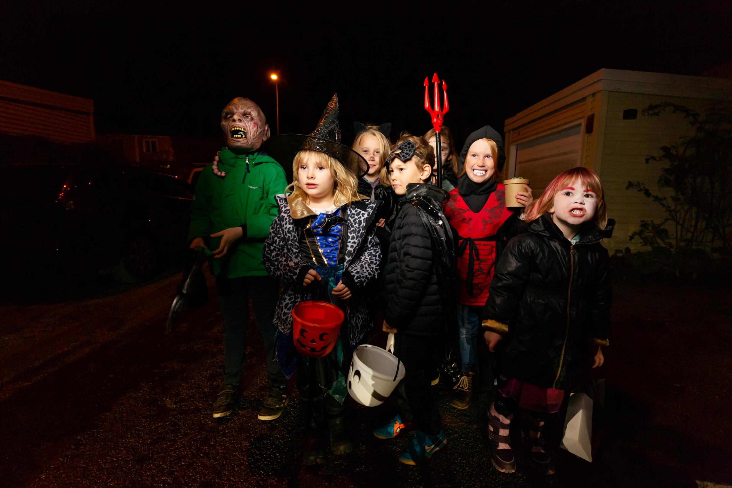 مجموعة من الأطفال يرتدون أزياء تنكرية احتفالصا بالهالوين و يسيرون في شارع مظلم ويجمعون الحلوى في أواعي بلاستيكية. 